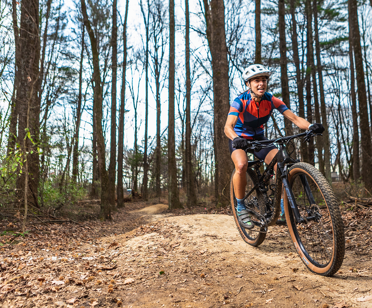 A biker wearing a helmet rides down a dirt trail in a stand of pine trees. 