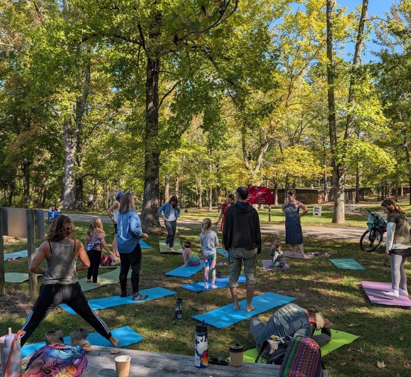 A group of families do yoga on blue mats underneath the shade of trees.