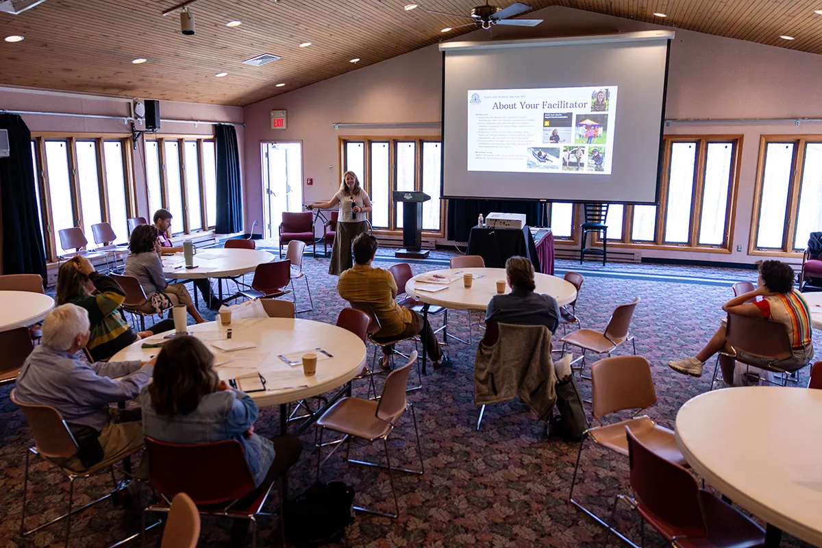 Presenter speaking to a small group seated at round tables during a workshop or training session in a conference room with a presentation displayed on a projection screen.