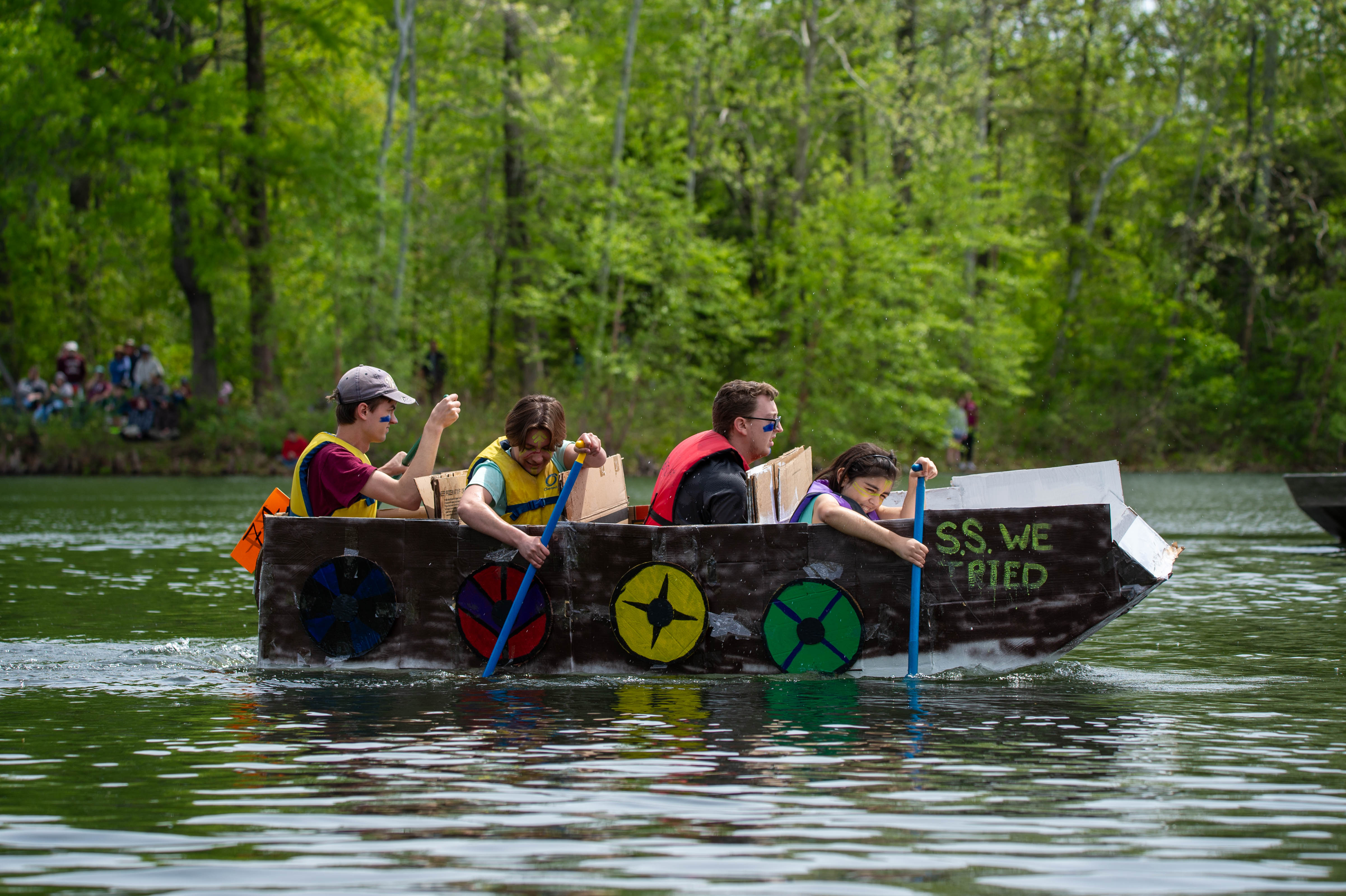 The image shows 4 students on a sunny day paddling a cardboard boat named the U.S.S. We Tried across Campus Lake.